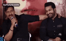 two men are sitting next to each other and one is singing into a microphone while the other is hugging him .