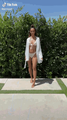 a woman in a bikini and a white robe is standing on a sidewalk in front of trees .