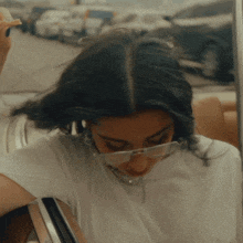 a woman wearing glasses and a white shirt is sitting in a car eating a sandwich .