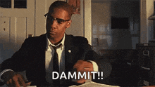 a man in a suit and tie is sitting at a table holding a book and saying `` dammit ! ''