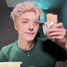 a young man in a green shirt is holding a burrito wrapped in aluminum foil