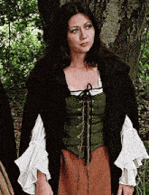 a woman wearing a green vest and a black cape is standing in a forest .