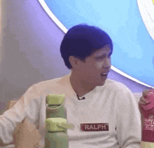 a man wearing a white sweater with the name ralph on it is sitting at a table holding a bottle .