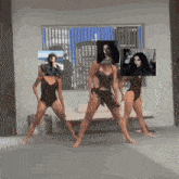 three women in black swimsuits are dancing in front of a window with a picture of a man on it
