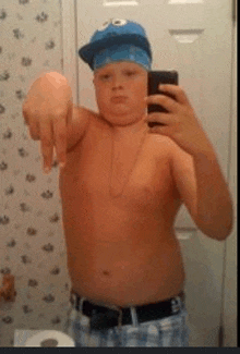 a shirtless boy wearing a cookie monster hat is taking a picture of himself in a bathroom mirror .