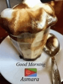 a cup of coffee with whipped cream and a spoon on a saucer with the words `` good morning asmara '' written on it .