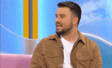 a man with a beard and a brown jacket is sitting on a pink couch talking on a television show .