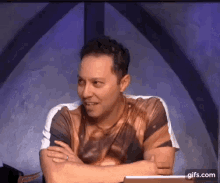a man is sitting at a table with his arms crossed and a t-shirt on .