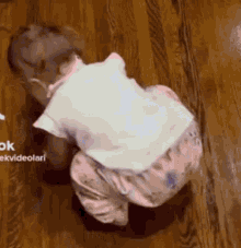 a baby is crawling on a wooden floor in a white shirt and pink pants .