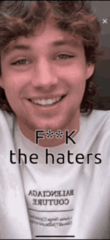 a young man with curly hair is smiling and wearing a white t-shirt that says `` f ** k the haters '' .
