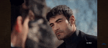 a man in a black turtleneck looks at another man in a black suit