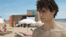 a young man with curly hair stands on a beach with umbrellas