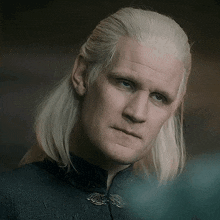a man with white hair and blue eyes is wearing a black shirt and a black jacket .