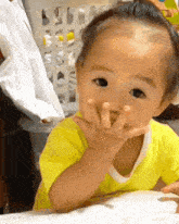 a baby in a yellow shirt is making a funny face with his hands .