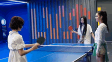 three women are playing ping pong in a room with a dart board in the background