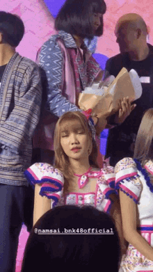 a woman holding a bouquet of flowers is surrounded by other people with the name namsai bnk48 official on the bottom right
