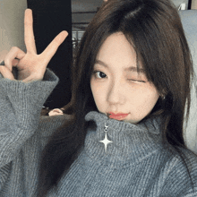 a girl wearing a grey sweater winks at the camera and makes a peace sign