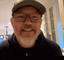 a man with a beard wearing glasses and a baseball cap is smiling .