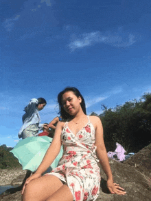 a woman in a floral dress sits on a rock with a green umbrella