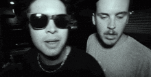 a man wearing sunglasses stands next to another man wearing a necklace