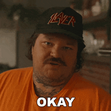 a man wearing a slayer hat says okay in an orange shirt