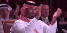 a man in a keffiyeh is sitting in a crowd of people and clapping .