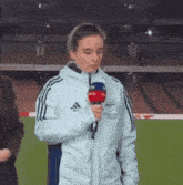 a woman in an adidas jacket is holding a microphone on a field