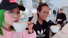two women are drinking coffee together in a cafe . one of the women has green hair .
