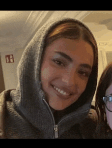 a woman wearing a hooded sweater smiles next to another woman wearing glasses