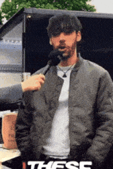 a man in a bomber jacket is being interviewed with a microphone and the word these is on the bottom right