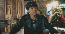 a young man wearing a leather jacket and hat is sitting at a table in front of a christmas tree