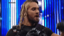 a man with long blonde hair is talking into a microphone while wearing a black shirt with the letter w on it .