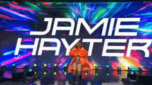 a woman in a fur coat is standing in front of a large screen that says jamie hayter