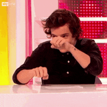 a man in a black shirt is sitting at a table covering his face with his hand .
