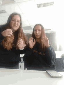two girls are posing for a picture in a classroom with a cell phone on the table between them .