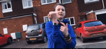 a young man wearing a blue sweatshirt is standing in front of a brick house holding a cell phone in his hand .