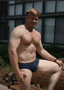 a shirtless man in blue swim trunks sits on a bench in front of a building