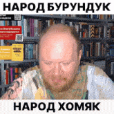 a bald man with a beard is sitting in front of a bookshelf with a yellow sign that says " kashina "