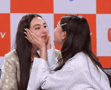 two women are touching each other 's faces in front of an orange and white checkered background with the letter v on it