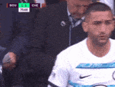 a man in a white nike shirt stands in front of a scoreboard that says bou 0-1 che