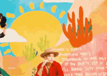a man in a cowboy hat is standing in front of a wall with a painting of a sun and cactus .