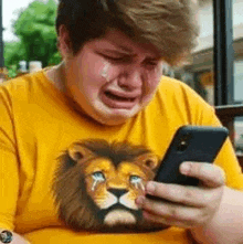 a man wearing a yellow shirt with a lion on it is crying while looking at his cell phone .