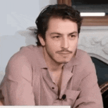 a man with a mustache is sitting at a table wearing a pink shirt and a microphone .