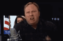 a man is talking into a microphone with the word you on the screen behind him