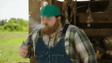 a bearded man wearing overalls and a green hat is blowing smoke out of his mouth .