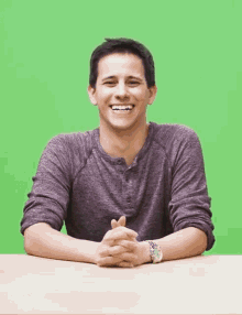 a man in a purple shirt sits at a table with his hands folded in front of a green screen