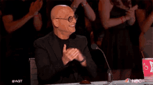 a man in a suit and glasses is sitting at a table clapping his hands in front of a microphone .