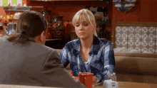 a woman in a plaid shirt sits at a table with a red mug