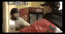 a man in a red jacket is talking to another man in front of a coca cola fridge