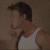 a man in a white tank top is standing in front of a wall and smoking a cigarette .